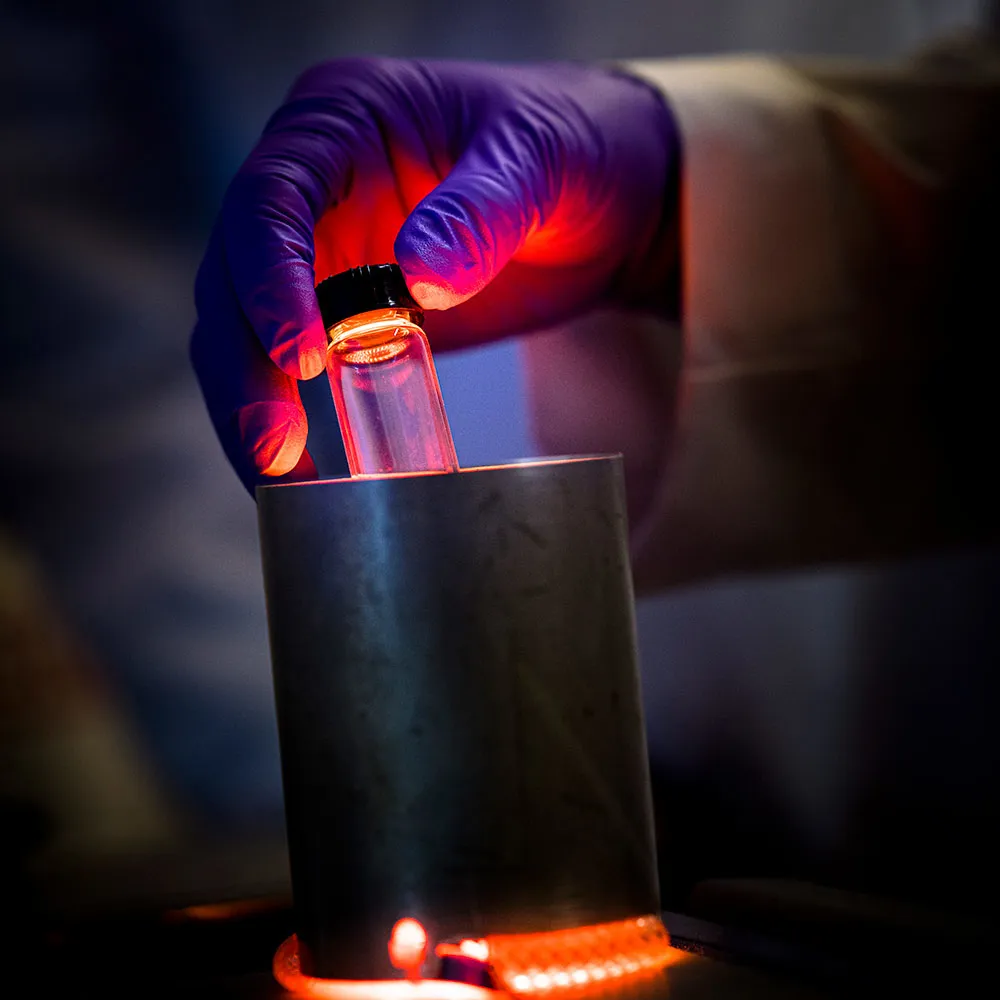 Gloved hand holding a test tube in a cylindrical container with an orange light illuminating the hand and test tube